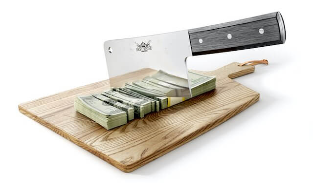 Stack of cash being cut into pieces by a meat cleaver on a wooden cutting board