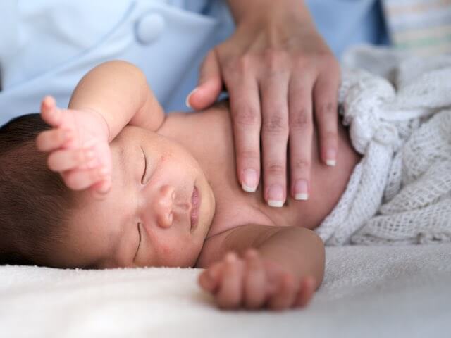 A mother's hand touches a baby lying sleeping on a bed