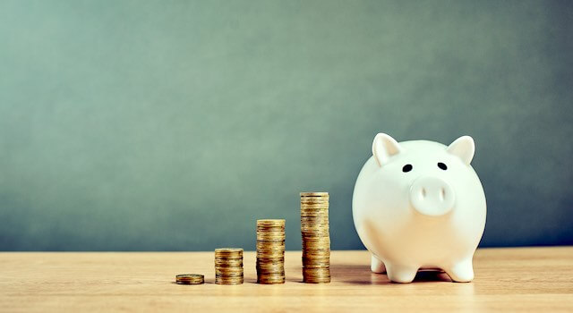 Four separate vertical stacks of coins growing in size from left to right pictured next to a white piggy bank on a wooden surface