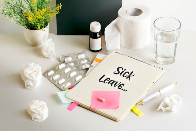 Words 'sick leave...' written on a small notepad on a desk next to used tissues, pills, a pen and a glass of water