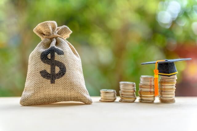 Burlap bag with a dollar sign on it next to four vertical stacks of coins growing in size from left to right with a student graduation cap on top of the last stack depicting student loan debt