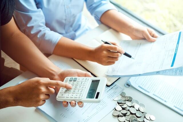 Man and woman sitting at a desk doing financial calculations; the woman is holding a notepad and pen and the man is performing a calculation with a calculator with coins and paperwork spread out in front of them