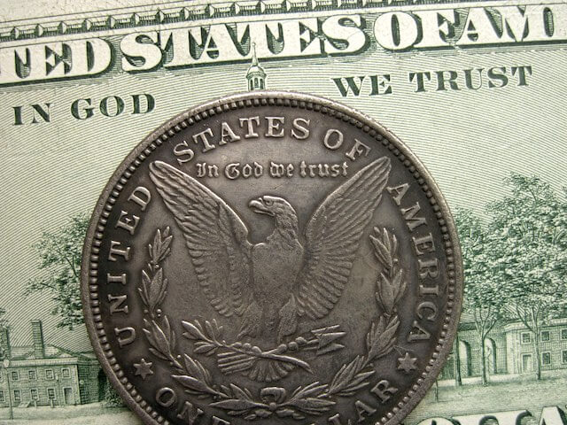 Close up of the words 'In God We Trust' on the back of a US dollar with a coin lying on top that also reads 'In God We Trust' with an eagle on it