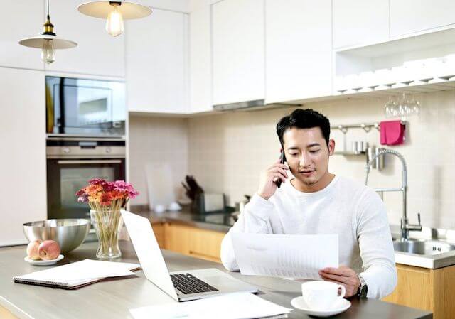 A millennial aged man is seen conducting telework in his kitchen as he talks on the phone and looks at a document with his laptop and a cup of coffee sitting on the counter in front of him