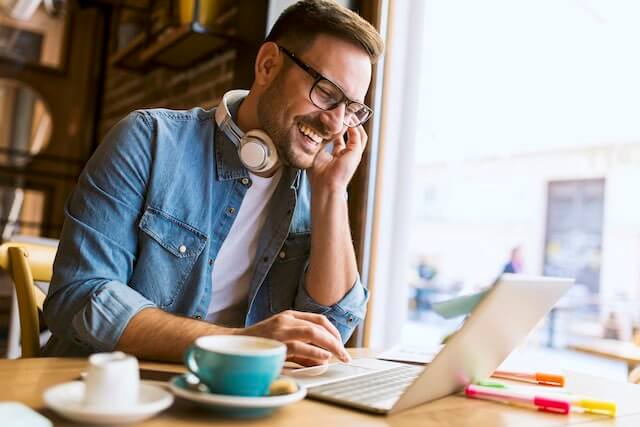 Man teleworking at a coffee shop - a man is seen smiling as he sits at a table working on his laptop with a cup of coffee on the table in front of him