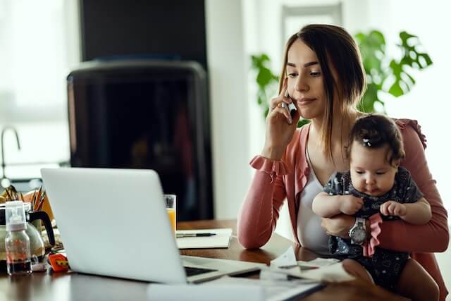 Young mother conducting telework at her home as she talks on the phone in front of a laptop computer while holding her baby
