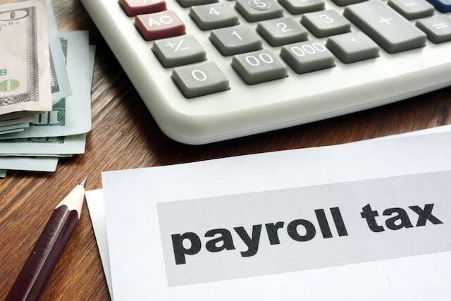 words 'payroll tax' on a sheet of paper on a desk next to a pen, cash and calculator