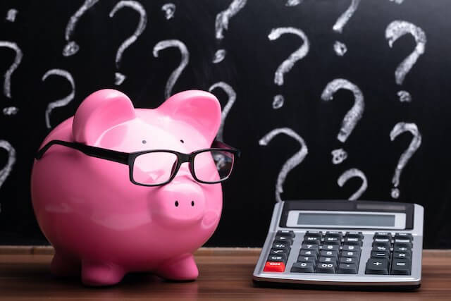 Pink piggy bank wearing glasses sitting on a desk next to a calculator with a bunch of questions marks drawn on a chalkboard in the background