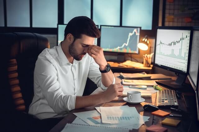 A man sits at his desk with his head in his hands appearing stressed out about his financial investments; financial graphs are pictured on the computer screens in front of him and there is paperwork on the desk