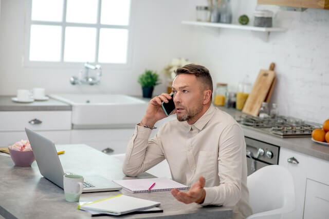 A man sits at a table in his kitchen conducting telework and talking on the phone and working in front of a laptop
