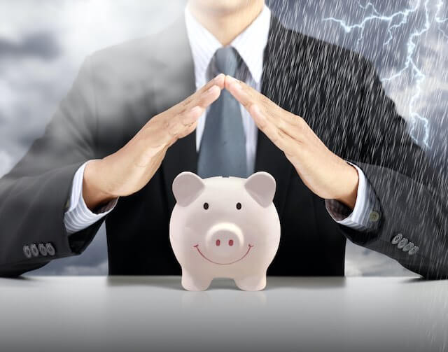 Businessman holding his hands in a v shape over the top of a smiling pink piggy bank to shield it from clouds, fog, and a thunderstorm depicting emergency savings or an emergency fund/rainy day fund