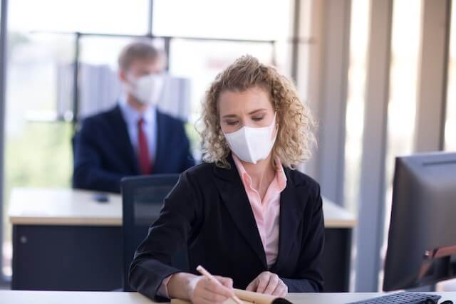 Young businesswoman wearing a surgical face mask amid the COVID-19 coronavirus pandemic while sitting at her desk in a corporate office writing on a note pad