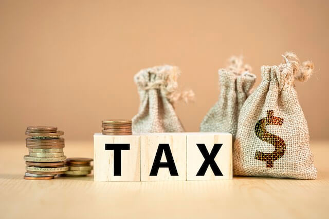 Words 'tax' written in wood block letters pictured in front of burlap bags of money and stacks of coins