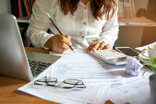 A woman sits in front of a laptop taking notes with a pencil as she pours over paperwork and spreadsheets doing accounting and financial planning