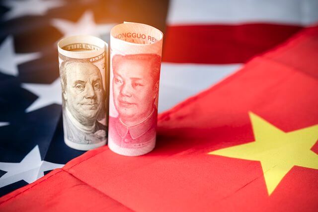 $100 bill rolled up to show the face of Benjamin Franklin and sitting up right vertically pictured right beside a Chinese Yuan rolled up; both bills are sitting on top of USA and China flags