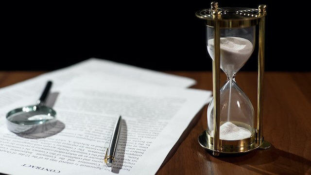 Contract document sitting on a desk next to an hourglass with a pen and magnifying glass on top of the contract papers