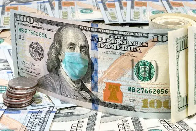 $100 bill pictured upright in a 3D fashion with the image of Benjamin Franklin wearing a surgical face mask to protect from COVID-19 coronavirus; a spread of $100 bills cash lies underneath the upright $100 bill