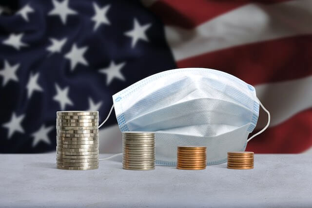 4 vertical stacks of coins decreasing in size from left to right pictured in front of a surgical face mask with a USA flag in the background