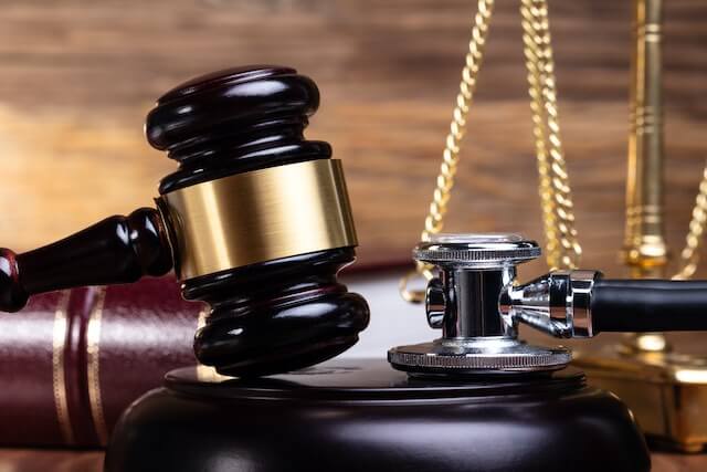 Wooden judge's gavel next to a stethoscope with a law book and justice scales pictured in the background