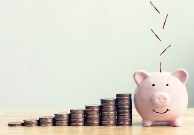 Vertical stacks of coins growing in height from left to right leading up to a pink piggy bank with coins falling into it from above depicting growing savings