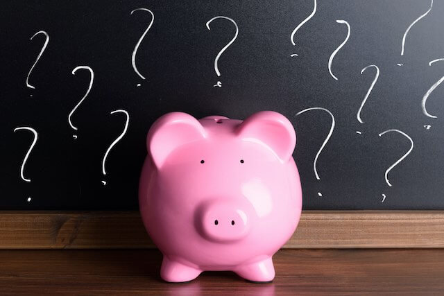 Pink piggy bank sitting on a wooden surface in front of a chalkboard with a series of question marks written on it