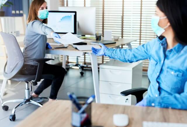 Two employees in an office practicing social distancing; they are wearing gloves and face masks and passing documents to each other while their desks are apart and facing in opposite directions