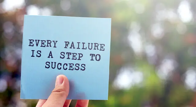 Close up of a person's hand holding an index card that reads 'every failure is a step to success'