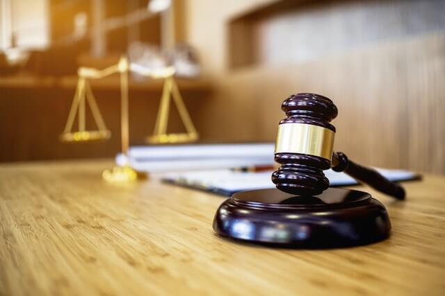 Wooden judge's gavel on a desk in the foreground with a notebook and justice scales pictured in the background