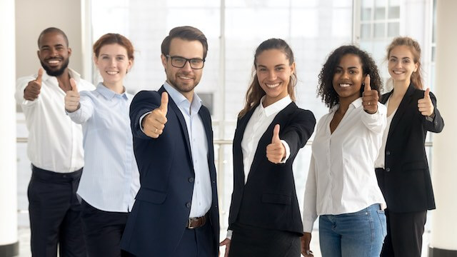 Group of 6 male and female employees smiling and giving a thumbs up sign