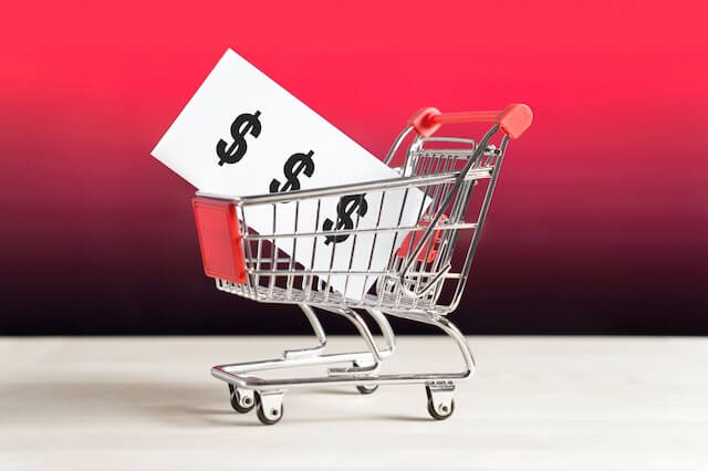 Grocery/retail store shopping cart with a white cardboard sign in it with three dollar signs ($) on it in black font