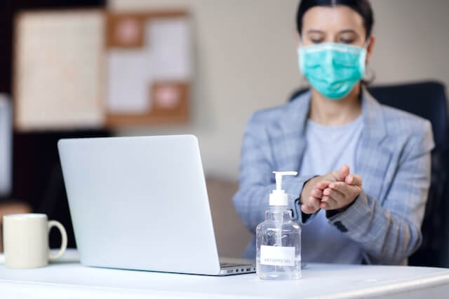 Woman sitting at her desk in front of a laptop wearing a face mask and applying hand sanitizer