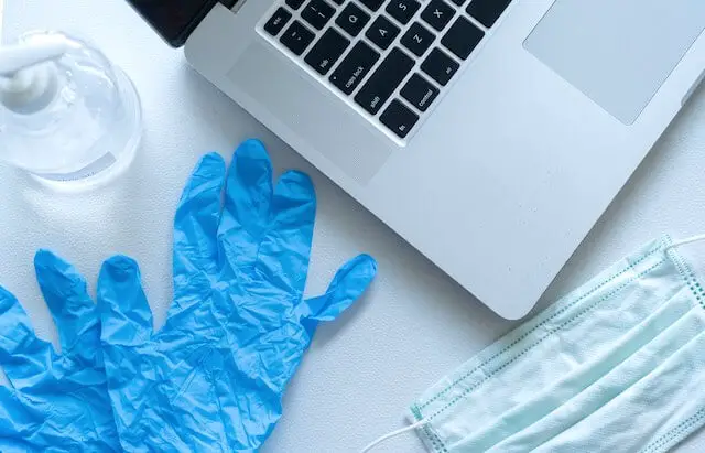 Hand sanitizer, disposable gloves, and a face mask sitting on a surface next to a laptop
