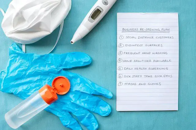 Digital thermometer, face mask, hand sanitizer and disposable gloves on a blue surface next to a note that is titled 'business reopening plan'