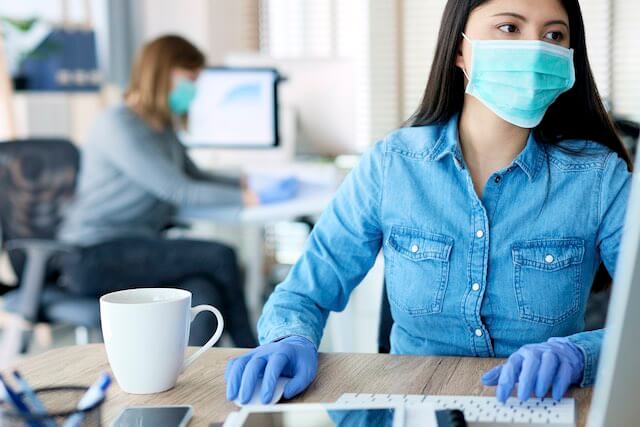 Woman sitting at her desk working on a computer wearing disposable gloves and a face mask