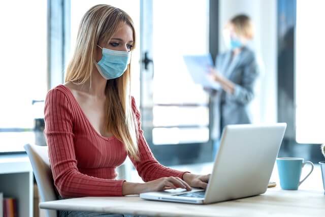 Female employee wearing a face mask sitting at her desk working on a laptop; another employee is pictured slightly blurred in the background wearing a mask