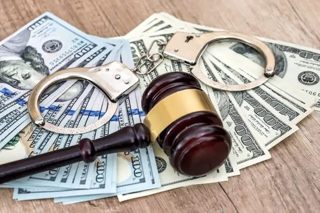 Wooden judge's gavel lying on top of a spread of $100 bills next to a pair of handcuffs on a wooden surface