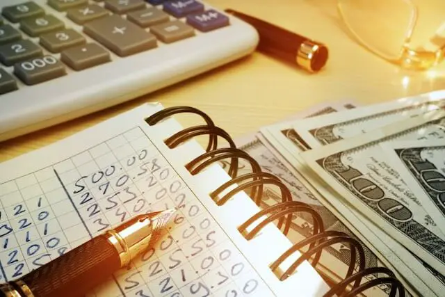 Notepad with columns of numbers written on it representing a budget next to a stack of cash and a calculator