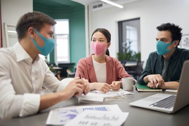 A group of three employees wearing face masks sitting around a table in an office having a meeting