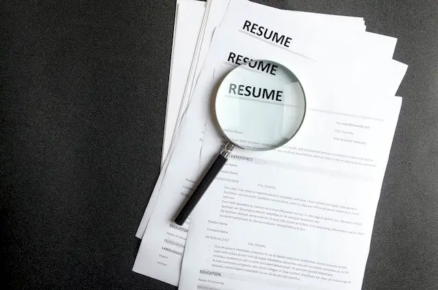Stack of paper resumes identified by the word 'resume' at the top of them with a magnifying glass sitting on top of them