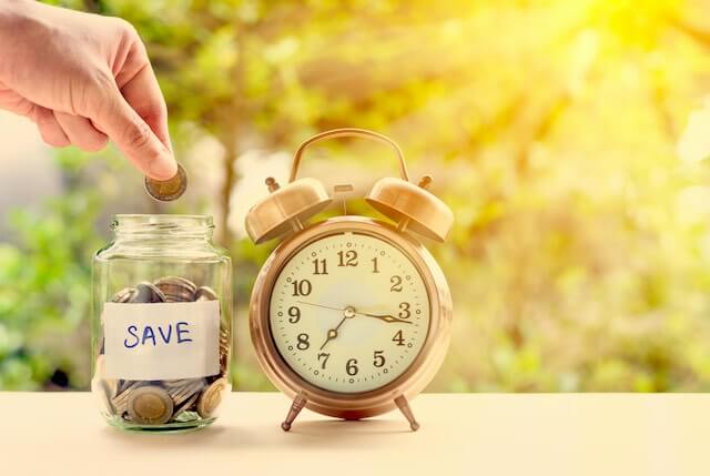 Close up of a person's hand putting money into a clear glass jar labeled 'save' next to an old style alarm clock