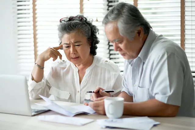 Senior aged Asian couple looking confused/frustrated as they sit in front of a laptop computer studying paperwork/bills