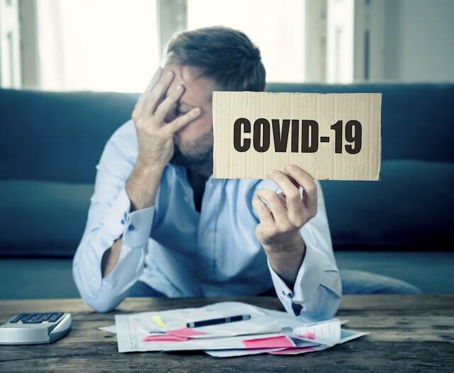 Man appearing frustrated/sad with his right hand over his face as he looks to the side while holding a sign that says 'COVID-19' in his left hand; a calculator and paperwork are on a table in front of him