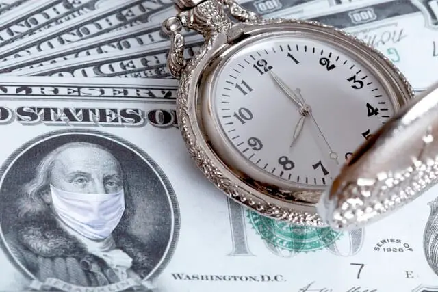 Close up of the face of a $100 bill with Benjamin Franklin pictured wearing a surgical face mask; a pocket watch sits on top of the $100 bill to the right of Franklin's photo
