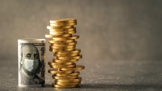 Rolled up $100 bill showing the face of Benjamin Franklin wearing a face mask standing upright next to a vertical stack of gold coins