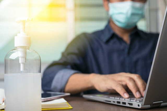 Employee working on a laptop wearing a face mask with a bottle of hand sanitizer in the foreground