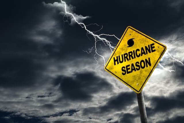 Storm clouds and a bolt of lightning pictured in the background behind a yellow road caution sign that reads 'hurricane season'