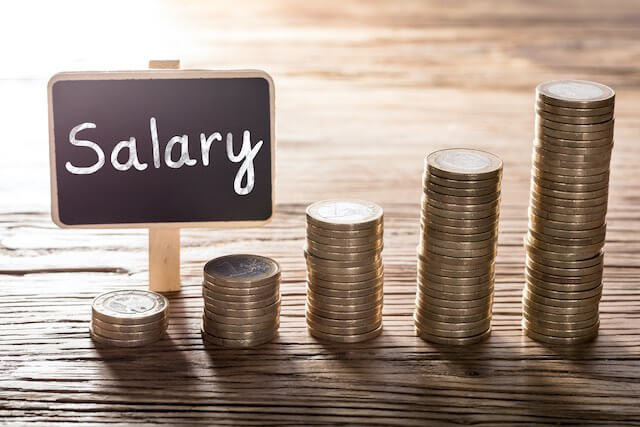 5 vertical stacks of coins on a wooden surface growing in height as they go from left to right with the word 'salary' written on a small chalkboard sign behind the stacks