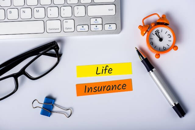 Two small strips of paper on a desk that read 'life insurance' surrounded by a paperclip, pen, reading glasses, keyboard and old style alarm clock