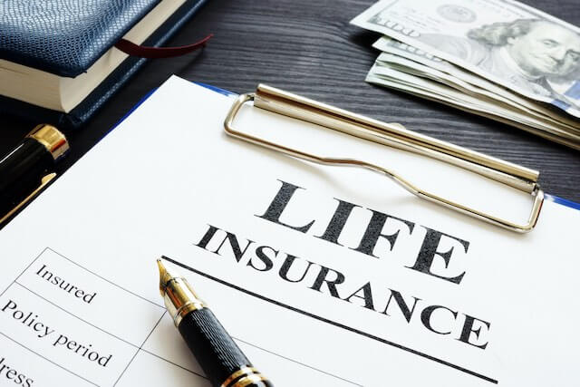 Words 'life insurance' printed at the top of a sheet of paper on a clipboard with a pen sitting on top of it; clipboard is lying on a desk next to a book and stack of cash