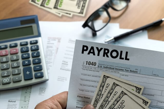 Close up of a person's hand holding a form labeled 'payroll' with some cash with a calculator, pen, glasses and other paperwork visible on the desk underneath